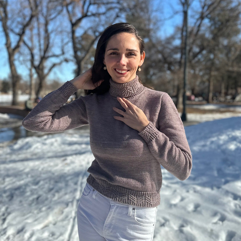 A white woman with brown hair is standing in a snowy park wearing a well fitting taupe turtleneck, and is holding her hair out of the way.